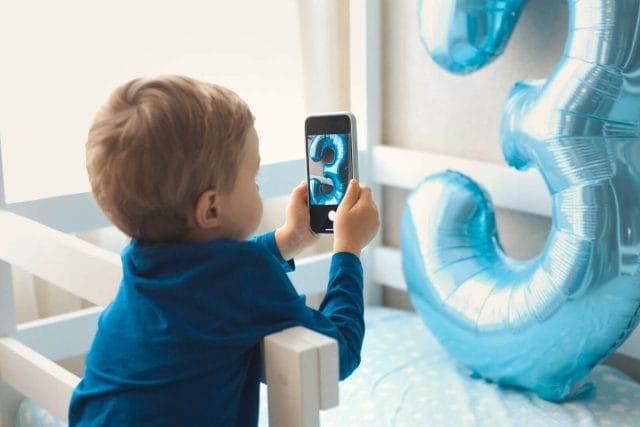 Little boy takes picture of number 3 balloon with smart phone.